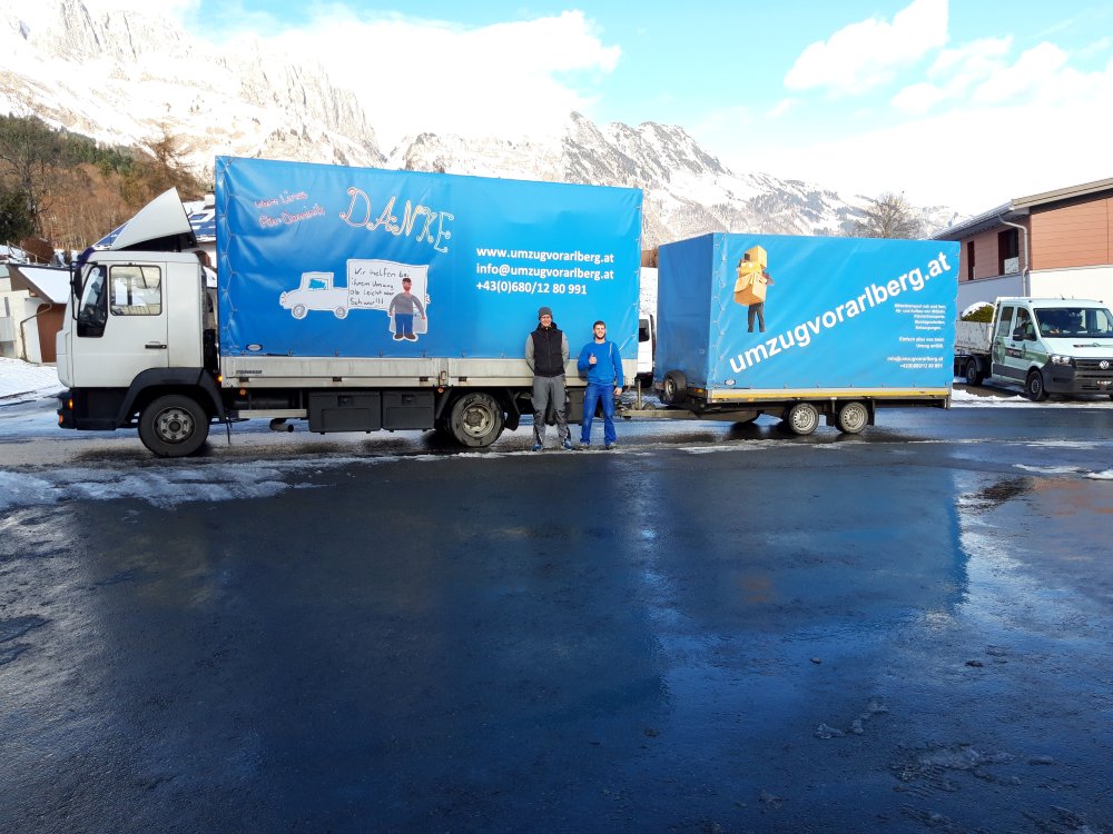 Zwei Umzugsarbeiter stehen vor zwei blauen Umzugsvorarlberg-LKWs, geparkt in einer verschneiten Berglandschaft.