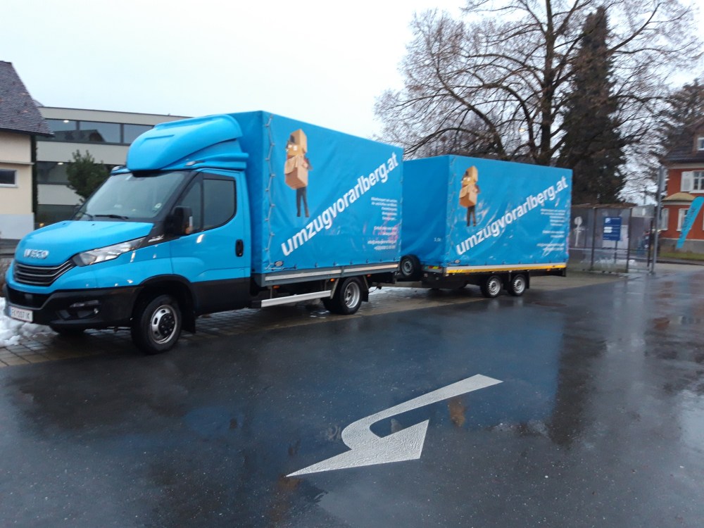 Ein blaues Umzugsvorarlberg-Transportfahrzeug mit Anhänger steht auf einer nassen Straße vor einem Gebäude.