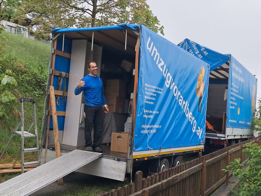 Ein Umzugsarbeiter steht lächelnd am LKW von Umzug Vorarlberg und zeigt den Daumen hoch, während Umzugskartons im Laderaum gestapelt sind.